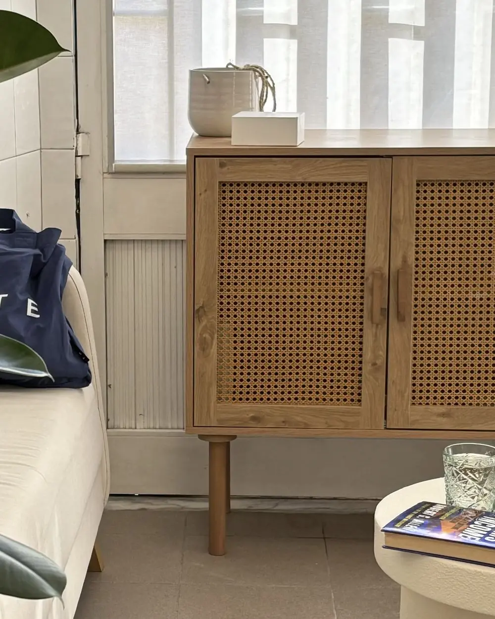 Le coin d’attente : un canapé clair, un buffet en cannage et la lumière filtrée par un store.
